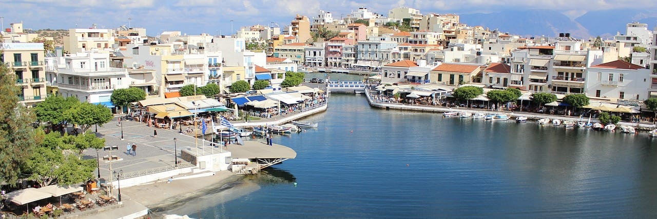 The circular inner harbour at Agios Nikolaos ringed by white and ochre buildings, fishing boats moored along the near quay and hills behind.