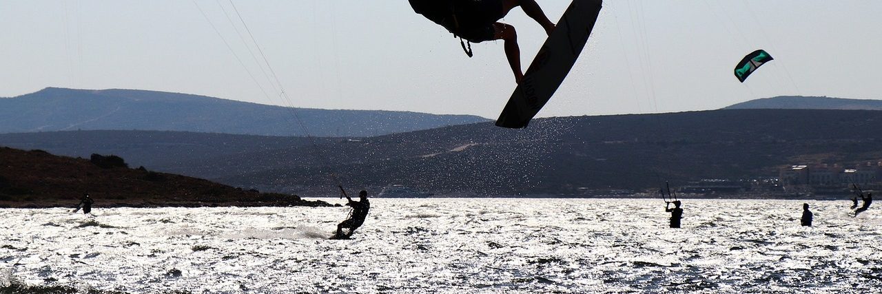 Kitesurfers on the glittering water of Alacati bay, one lifted clear of the surface, with the low hills of the Cesme peninsula behind them.