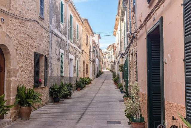 A narrow lane of shuttered stone houses in Alcudia old town, potted plants along the walls.