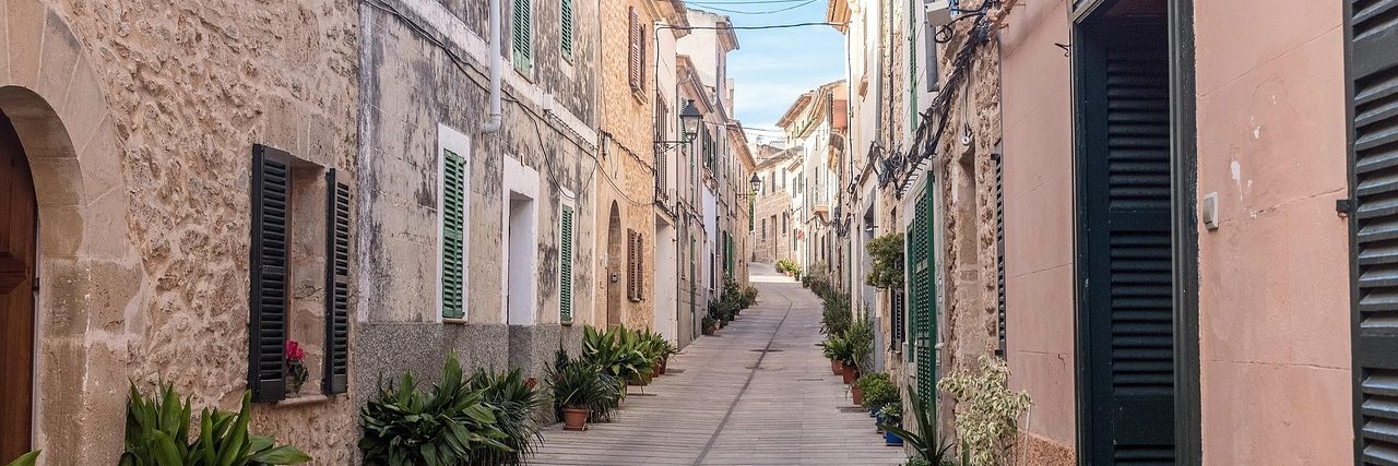 A narrow lane in Alcudia old town, sandstone and painted houses on both sides with green louvred shutters, potted plants along the walls and no traffic.