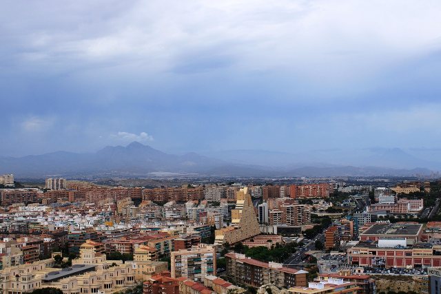 Dense apartment blocks and tiled rooftops of Alicante, with wooded hills and a hazy mountain ridge behind them.
