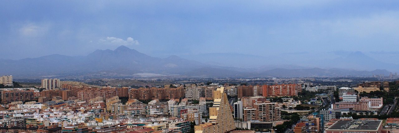 Alicante seen from above, apartment blocks and tiled roofs spreading inland towards a ridge of hazy mountains under heavy cloud.