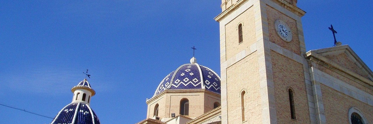 The blue and white tiled domes of Altea's church beside its stone bell tower and clock, against a deep blue sky.