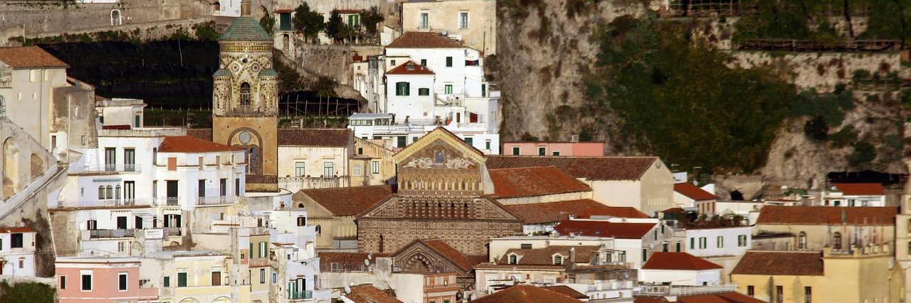 Amalfi packed tightly up the cliff behind its seafront, pale houses stacked in terraces with the cathedral's bell tower rising among them and bare rock above.