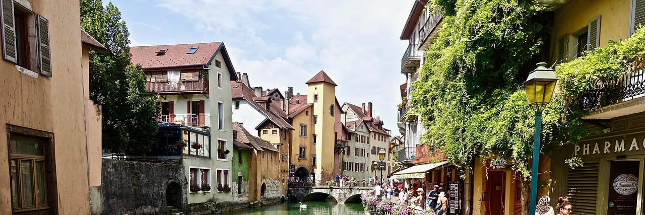 A green canal in the old town of Annecy running between old houses, flowers along the railings and an arched bridge at the far end.