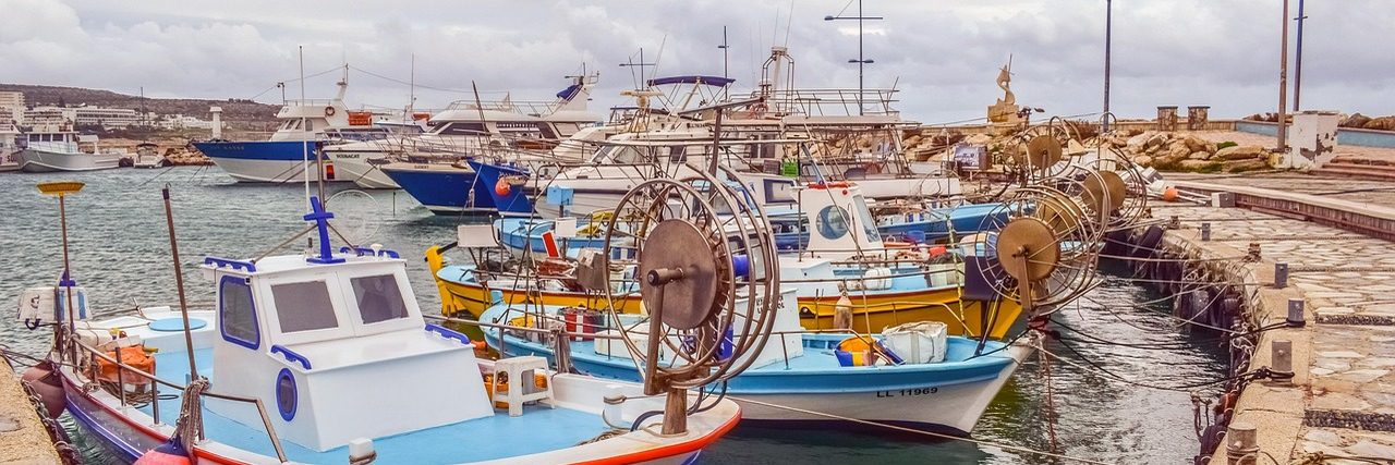 The fishing harbour at Ayia Napa, packed with small wooden boats painted blue, yellow and red, larger craft moored behind them and a stone quay running along the right.