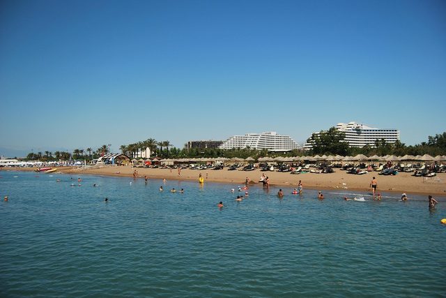 A sandy beach at Belek with bathers and low hotels among pines behind.