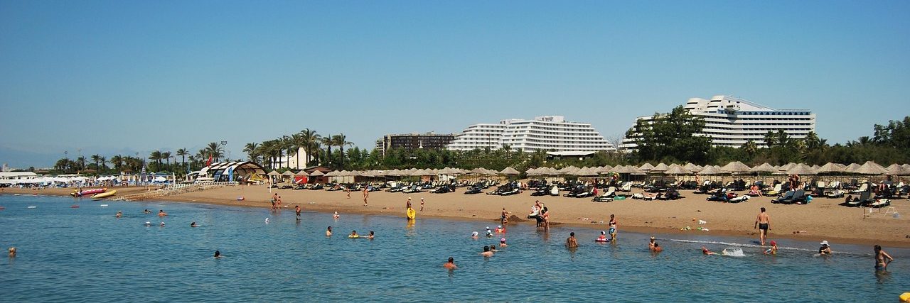 A long sandy beach at Belek with bathers in shallow water, and low hotel buildings set back among pine trees along the shore.