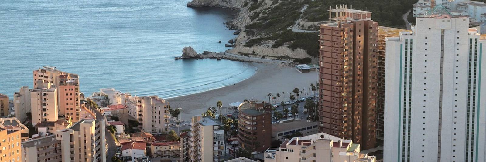 Benidorm from above, apartment blocks and hotel towers packed along a hillside with a curved sandy cove and turquoise water beyond them.