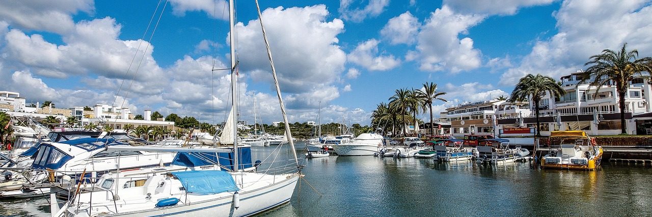 Yachts and motor boats moored along both sides of the marina inlet at Cala d'Or, white low-rise buildings and palms behind them under a sky of broken cloud.
