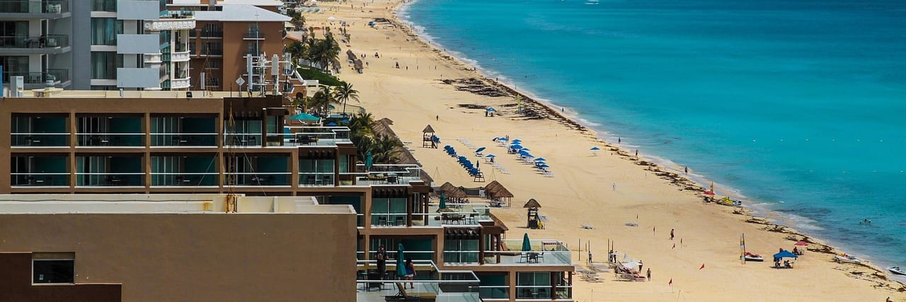 A long beach in the Cancun hotel zone curving away between high-rise hotel blocks and turquoise sea, a hotel balcony in the near foreground.