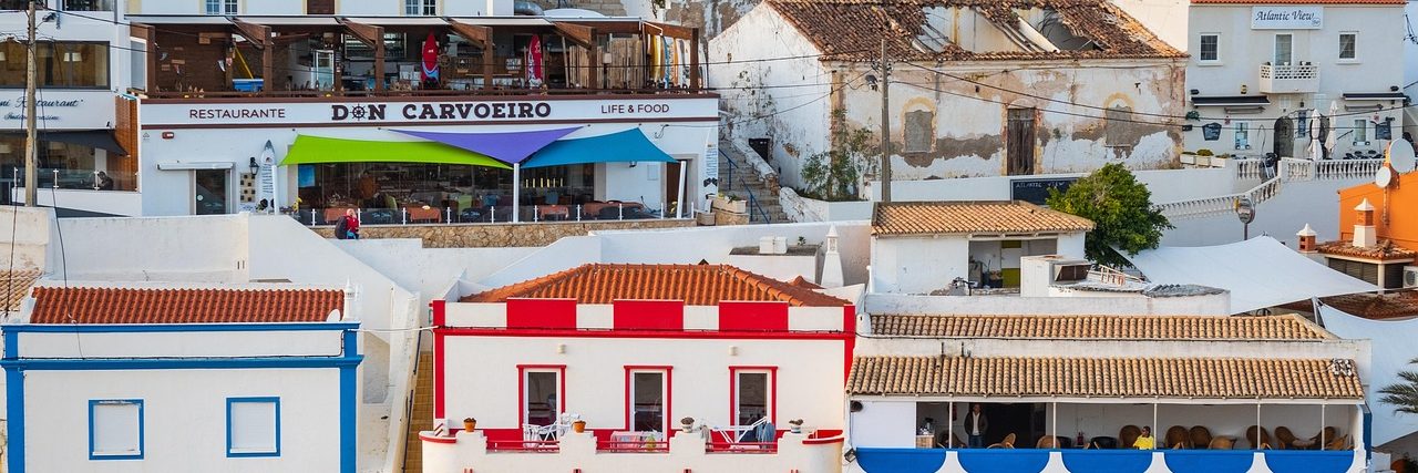 The square behind Carvoeiro's beach, white houses climbing the slope above two buildings painted in blue and white and in red and white stripes.
