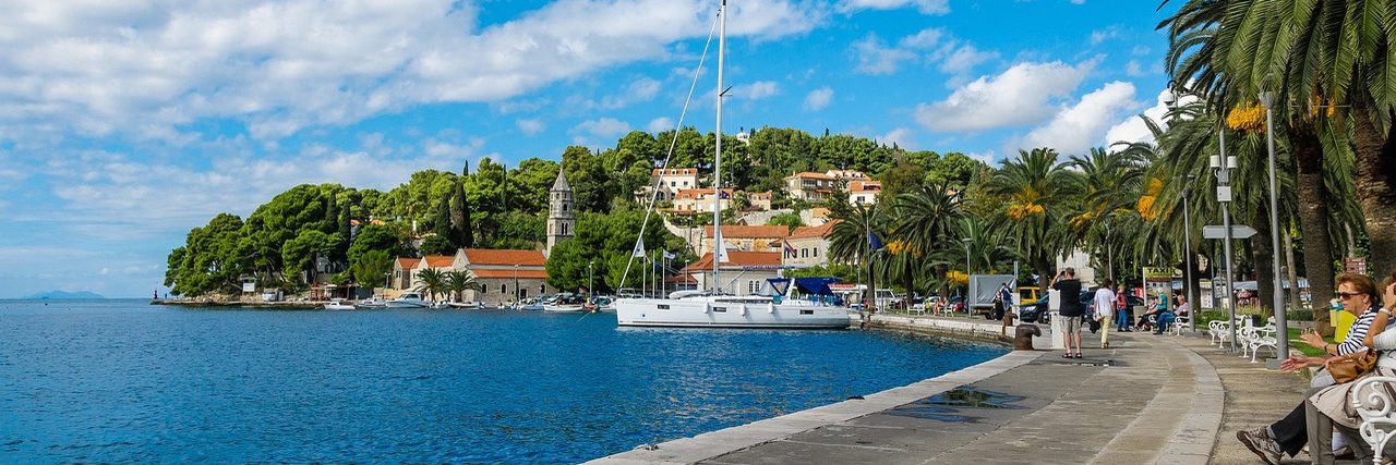 The quay at Cavtat with a yacht moored alongside and people sitting under palms, and across the water the wooded peninsula with a stone church tower and red-roofed houses among the pines.