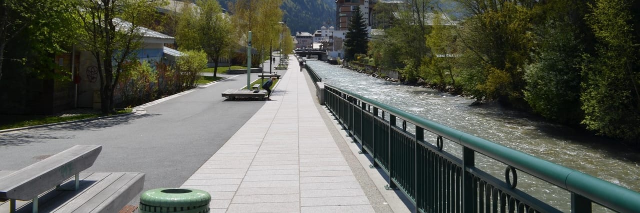 A paved riverside promenade in Chamonix with a metal railing and bare trees, the snow-covered Mont Blanc massif filling the head of the valley.