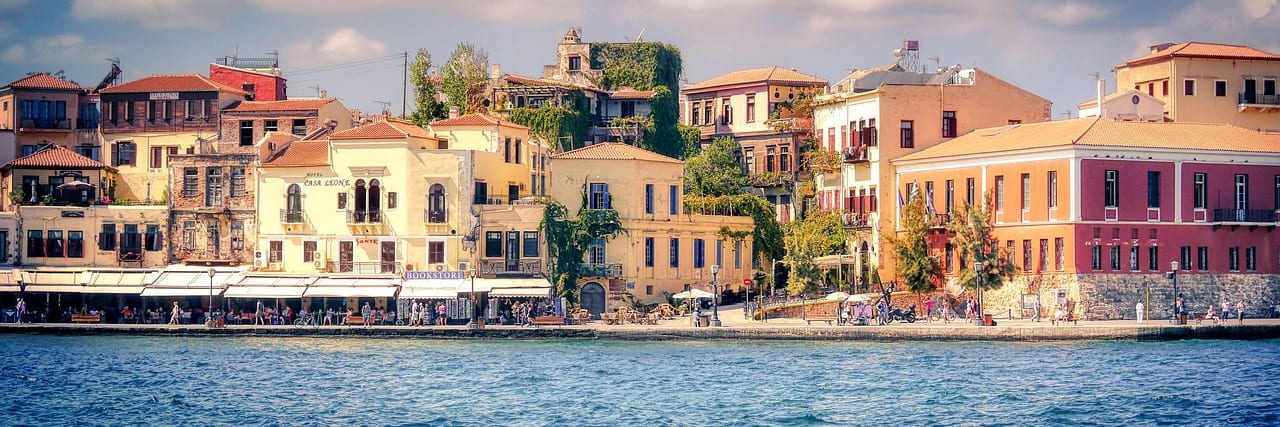 A row of tall painted houses along the Venetian waterfront at Chania seen across the harbour water, hills rising behind the town.