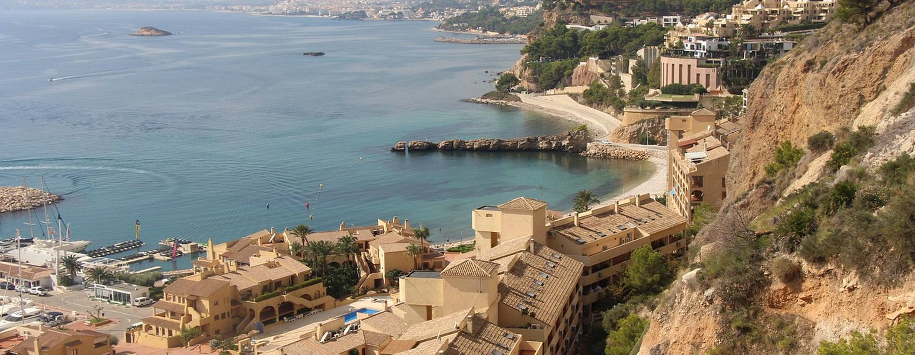 A bay on the Costa Blanca from above, turquoise shallows and a small harbour with low sandstone buildings stepping down a wooded headland.