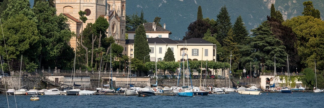 A bell tower and church among trees on the shore of Lake Como, villas along the water and moored sailing boats in front, wooded mountains rising behind.