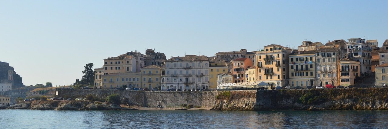 The Venetian seafront of Corfu Town seen from the water: a row of tall ochre, cream and pink houses standing along the top of a stone sea wall, with cars parked on the road beneath them and the old fortress at the left edge.