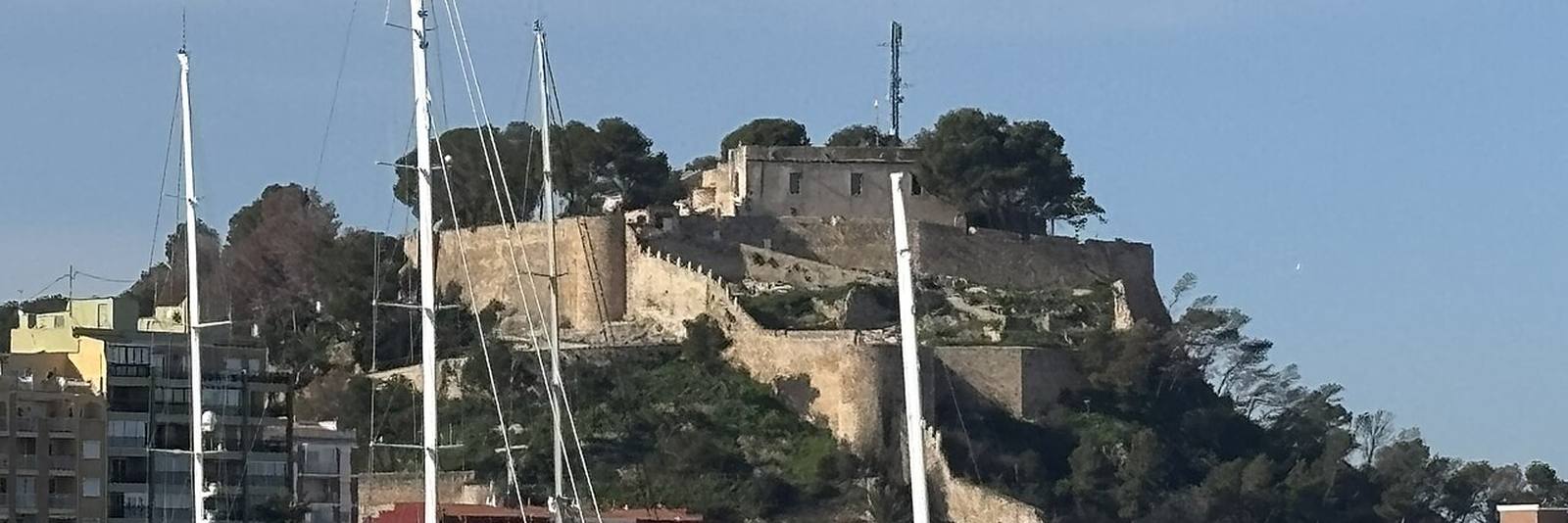 The castle at Denia on its wooded hill, seen past the masts of yachts moored in the marina below.
