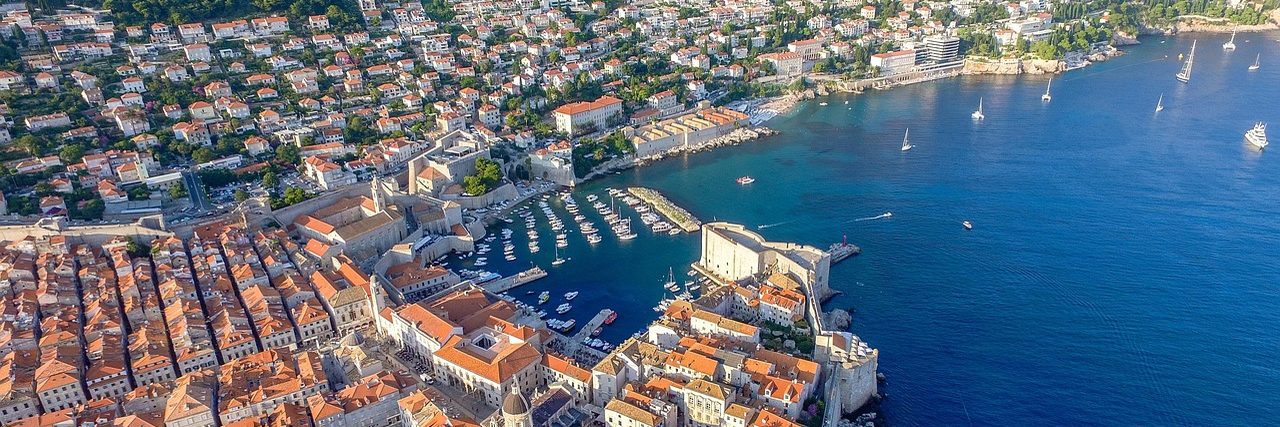 Dubrovnik's walled old town from the air: densely packed terracotta roofs running down to the old port, boats moored inside the harbour mole and open blue sea beyond.