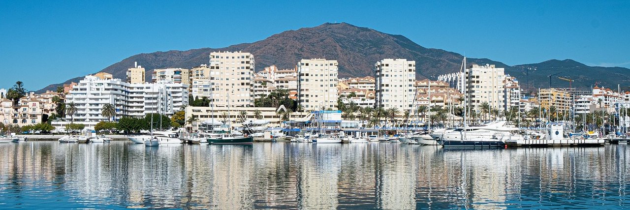 Estepona seen across still harbour water, white apartment blocks and moored yachts reflected below a bare mountain.