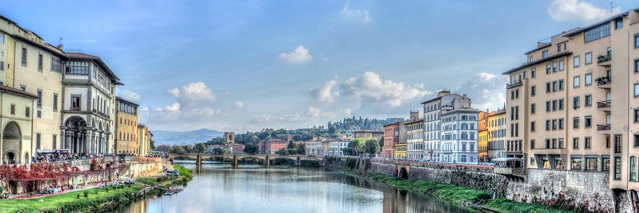 The Arno running between its embankments in Florence, ochre and cream buildings along both banks and trees overhanging the water on the near side.