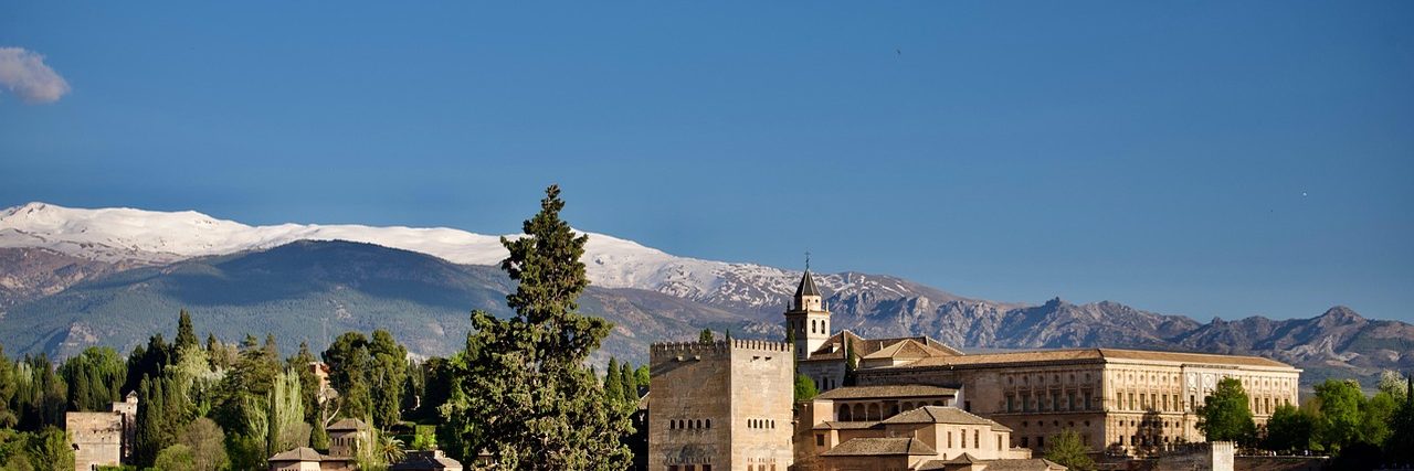 The Alhambra along its ridge at Granada: a square battlemented tower and the long pale facade of the palace among dark cypresses, with the snow-covered Sierra Nevada rising behind them under a clear sky.