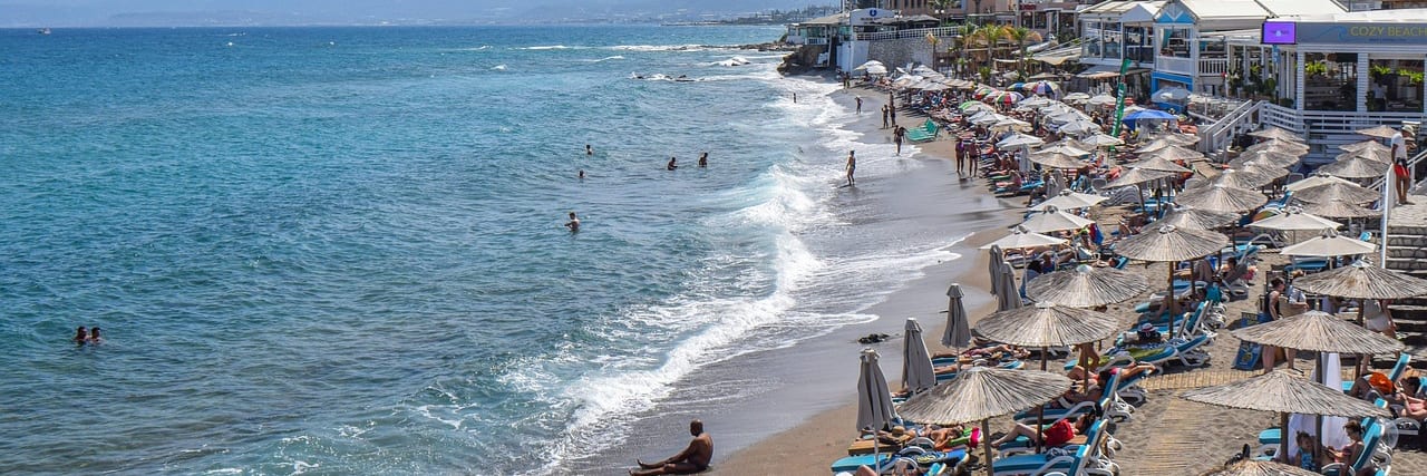 A curving beach at Hersonissos lined with sunbeds and parasols, swimmers in shallow turquoise water and the resort's buildings along the shore.