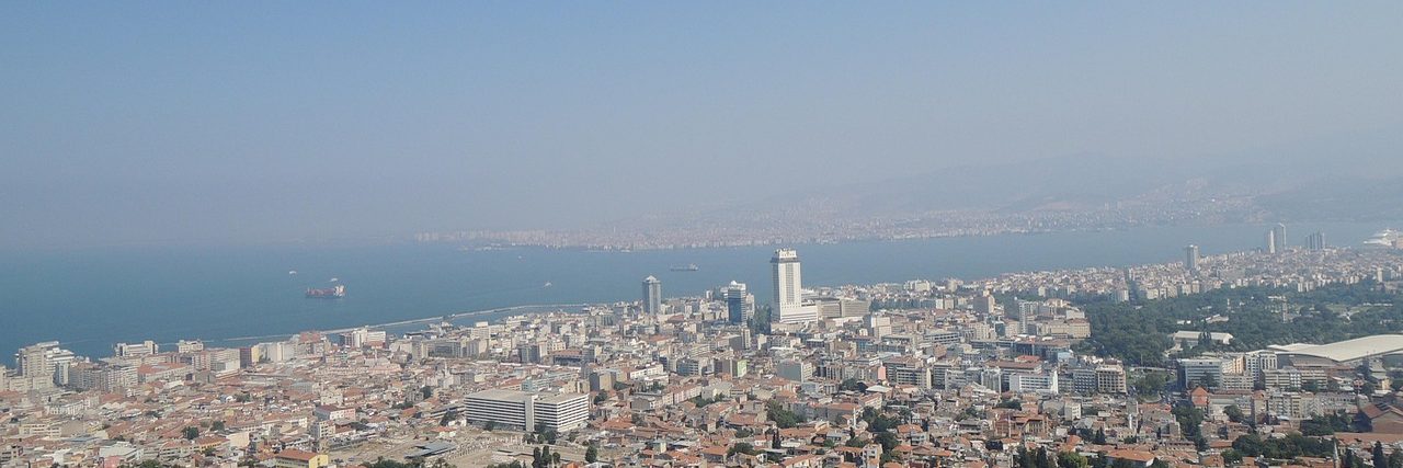 Izmir seen from the hills above it: rooftops in the foreground giving way to tower blocks and the long curve of the bay fading into haze.