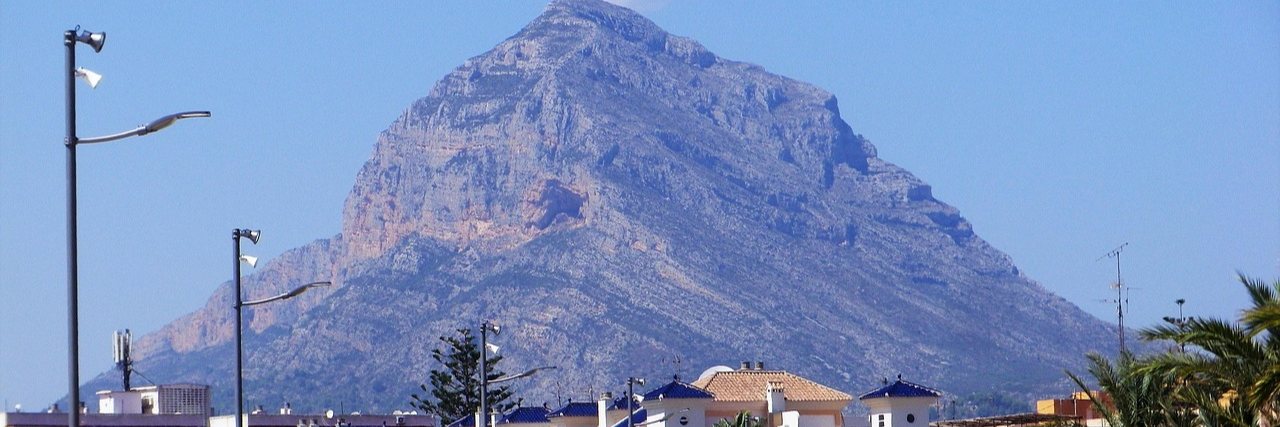 The bare rock of the Montgo massif rising steeply behind Javea, with blue-tiled roofs, street lights and a palm along the bottom of the frame.