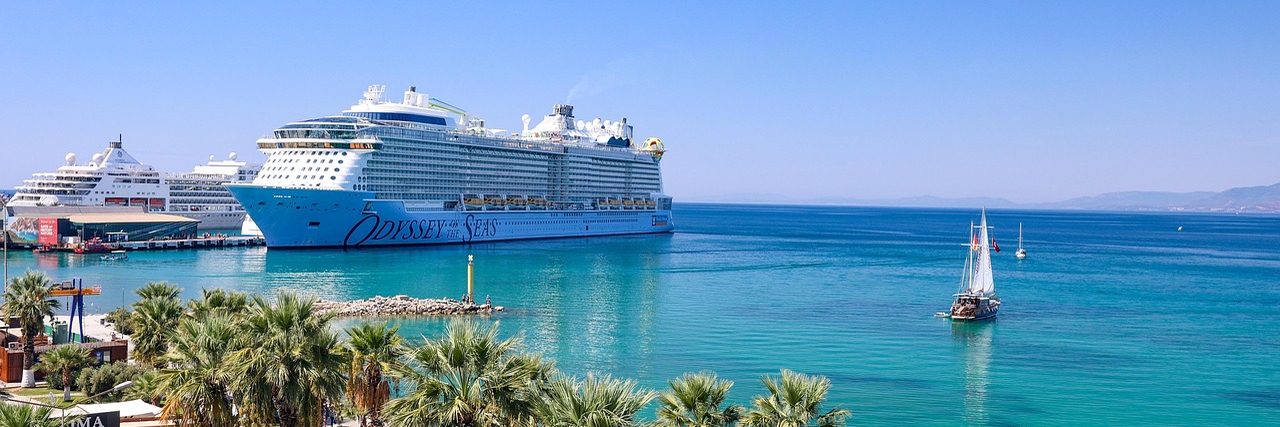 A large cruise ship berthed at Kusadasi with a second one beyond it, a sailing boat out on turquoise water and a line of palm trees along the front.