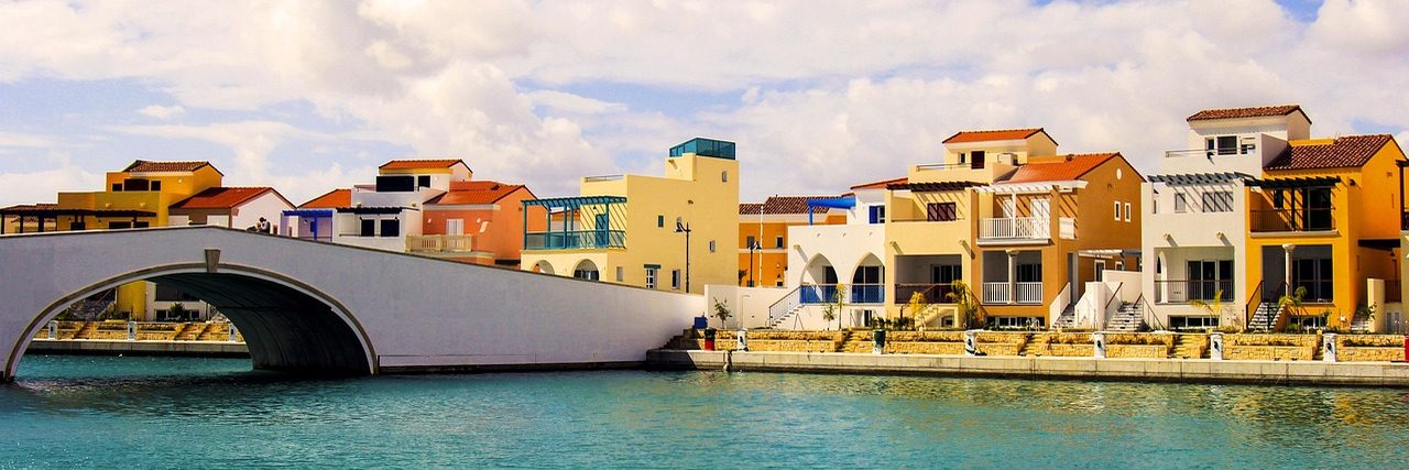 Limassol Marina: a white pedestrian bridge arching over the channel, with terraces of ochre and cream waterfront houses along the quay behind it and calm water in front.