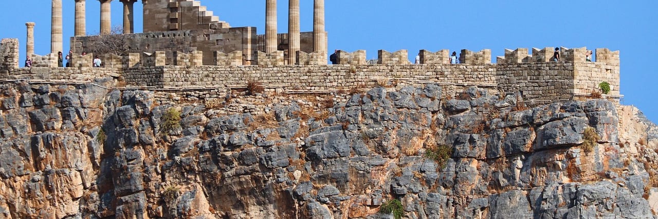 A colonnade of standing columns and ruined walls along the top of a sheer rock cliff at Lindos, plain blue sky behind them.