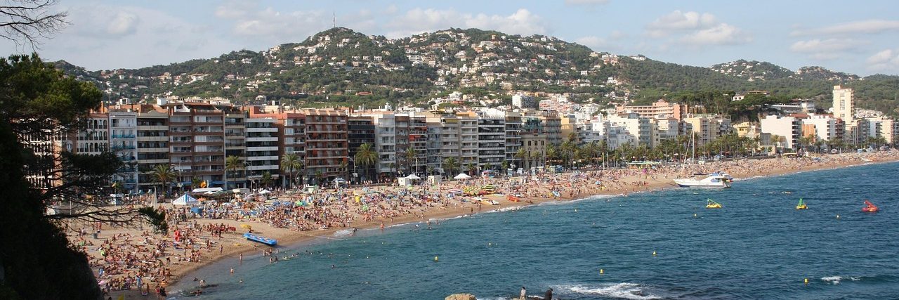 The crowded main beach at Lloret de Mar curving away to a rocky pine-covered headland, with the town's white blocks behind the sand.