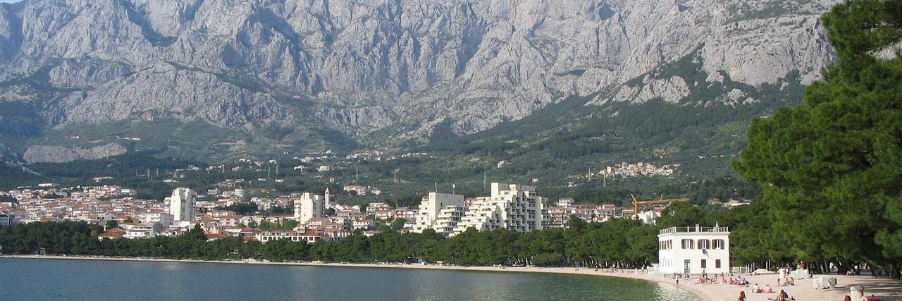 The grey limestone wall of the Biokovo massif rising behind Makarska, the white town spread along the shore beneath it and a curve of pebble beach with bathers in the foreground.
