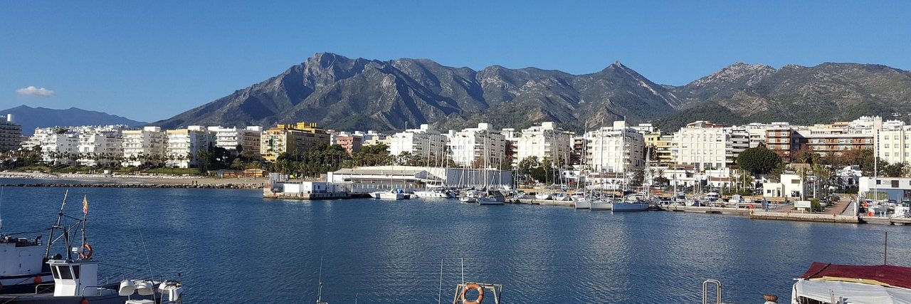 Fishing boats moored in Marbella harbour, white apartment blocks along the front behind them and the bare ridge of La Concha rising over the town.
