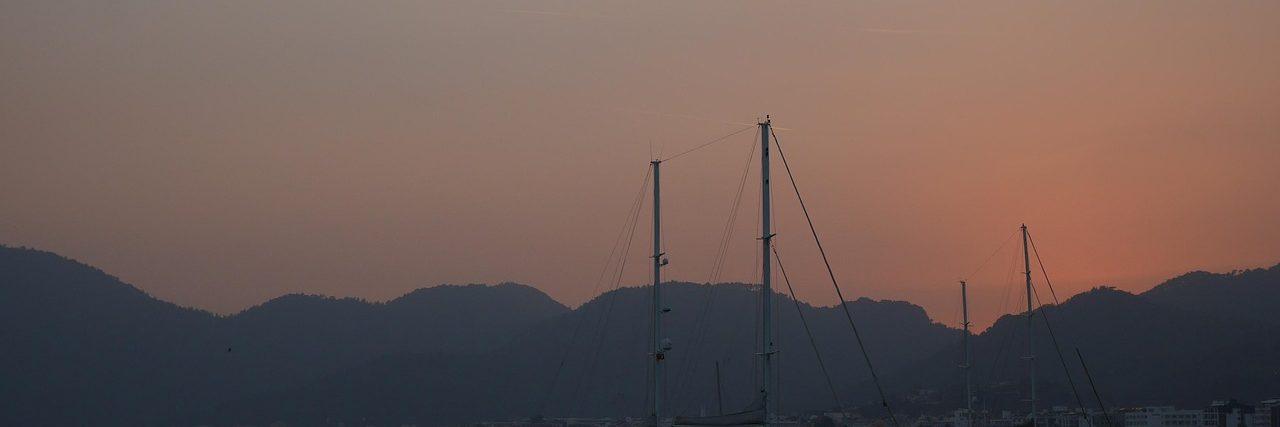 Traditional wooden gulets lying at anchor off Marmaris at dusk, their masts bare against a pink sky and the mountains of the gulf behind them.