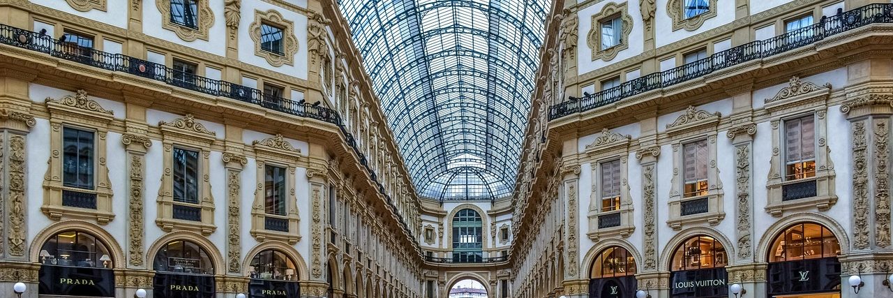 The Galleria Vittorio Emanuele II in Milan looking down the arcade, its glass and iron vault overhead, painted lunettes above the shopfronts and the mosaic floor below.