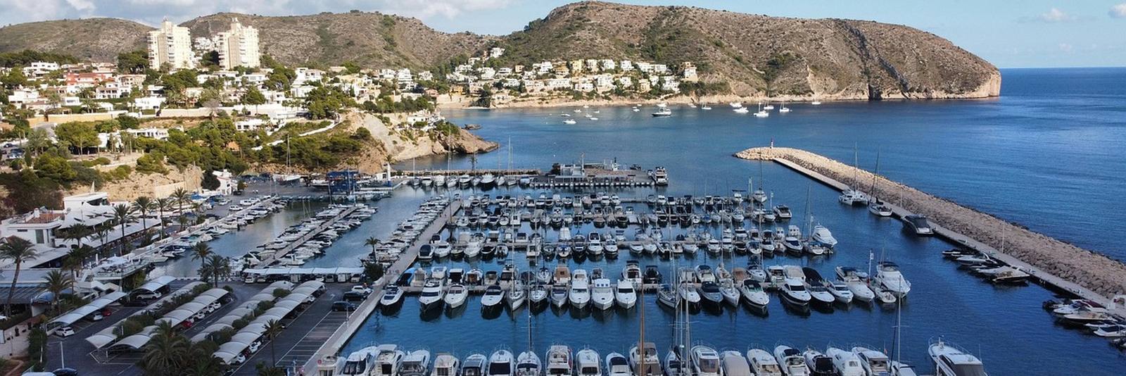 The marina at Moraira from above, rows of white boats behind a long breakwater, the town on wooded slopes at the left and a headland closing the bay at the right.