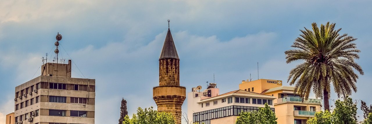 Rooftops in Nicosia: a concrete office block on the left, a stone minaret rising in the middle, low modern buildings behind and a tall palm on the right.