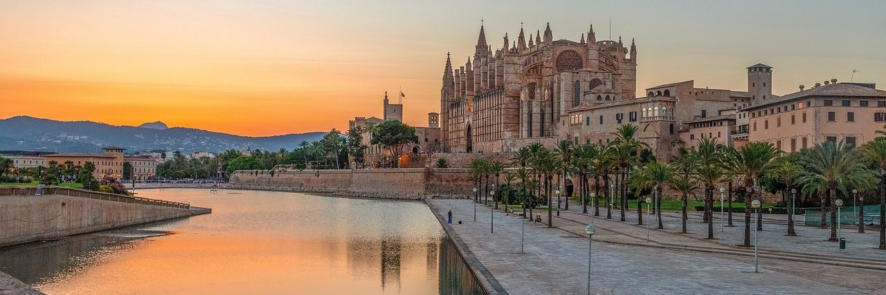 Palma cathedral standing above the old sea wall at sunset, its west front lit orange, with the Parc de la Mar water reflecting it and a palm-lined promenade running along the near side.