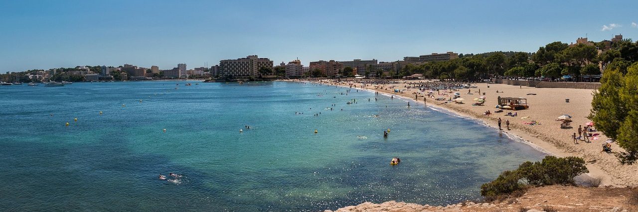 The shallow turquoise bay at Palma Nova seen from the rocks at one end, a curve of sand with sunbathers and low hotel blocks along the back of it.