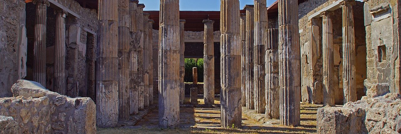 A colonnade of brick and stone columns around a roofless courtyard at Pompeii, the walls standing to about head height and grass growing over the floor.