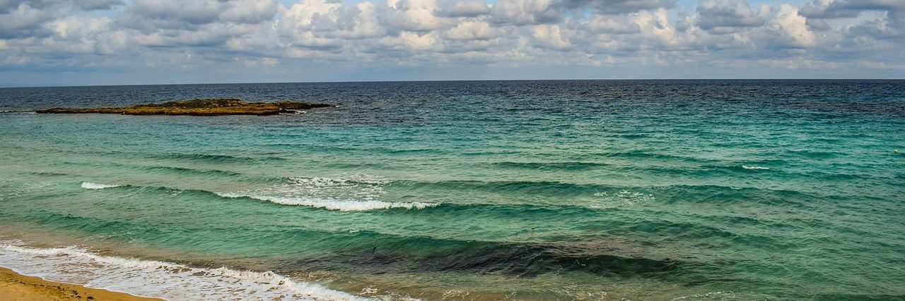 Fig Tree Bay at Protaras: a strip of pale sand in the foreground, clear turquoise shallows darkening to blue, the low rocky islet that sits off the beach, and a bank of cloud on the horizon.