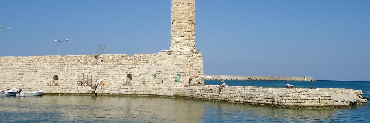 A stone lighthouse at the end of the harbour mole at Rethymno, small boats moored along the wall and open sea beyond.