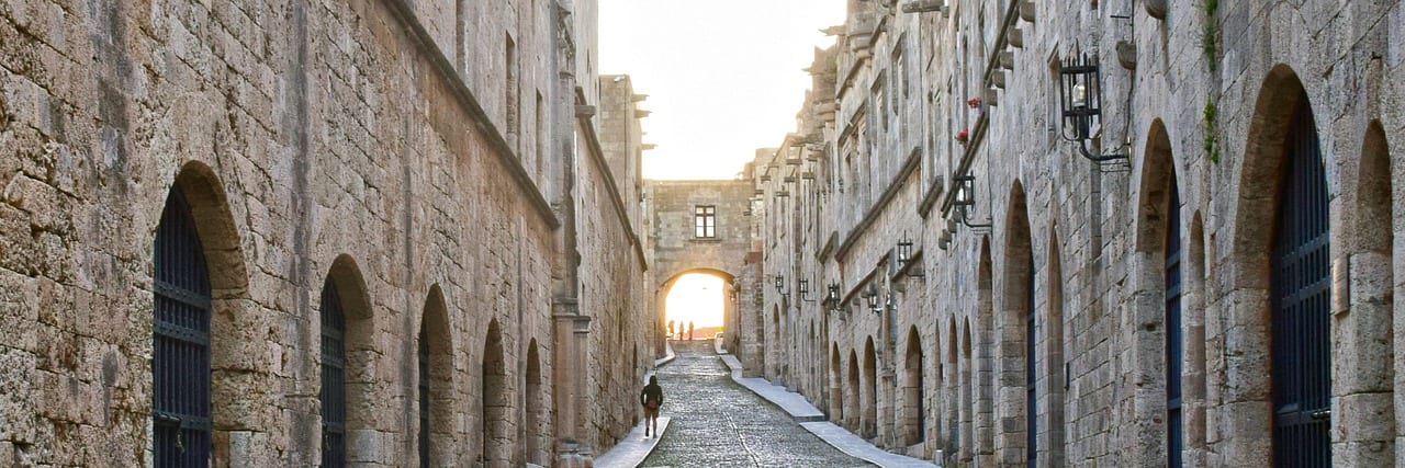 A narrow cobbled street in Rhodes old town between two ranks of arched stone buildings, rising to an archway at the far end with one figure walking under it.