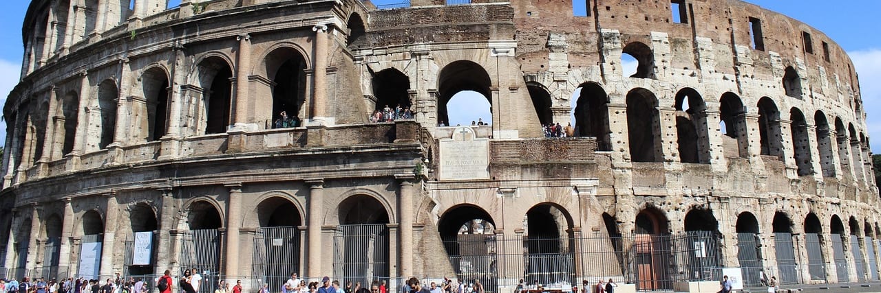The outer wall of the Colosseum in Rome, two tiers of arches in weathered stone, with people along the railing at street level.