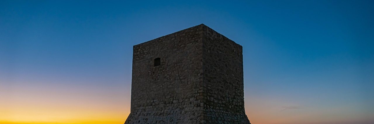 A square stone watchtower standing alone on a low spit in the Santa Pola salt lagoon, its walls catching the last light against a graded dusk sky.