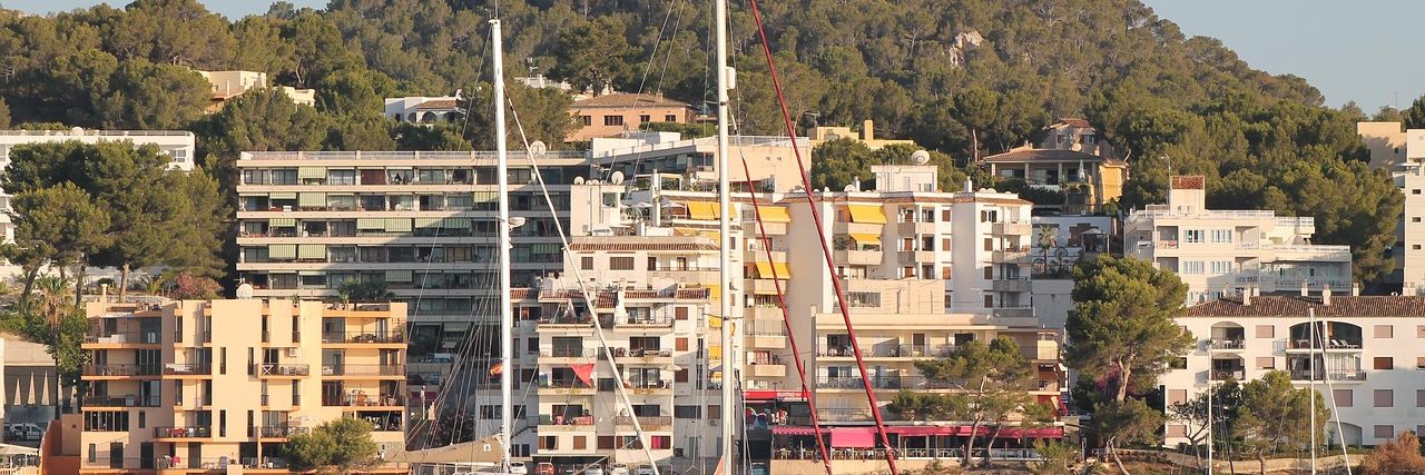 Apartment blocks and low white buildings stepping up a pine-covered hillside above Santa Ponsa, with the masts of moored yachts rising in front of them.