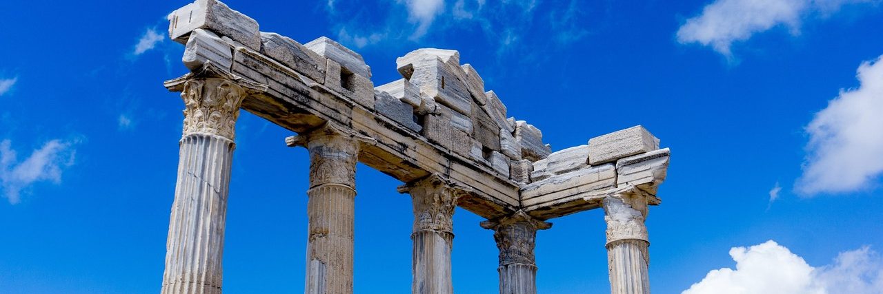 Three standing columns of the ancient temple at Side carrying a fragment of entablature, seen from below against a blue sky with scattered cloud.